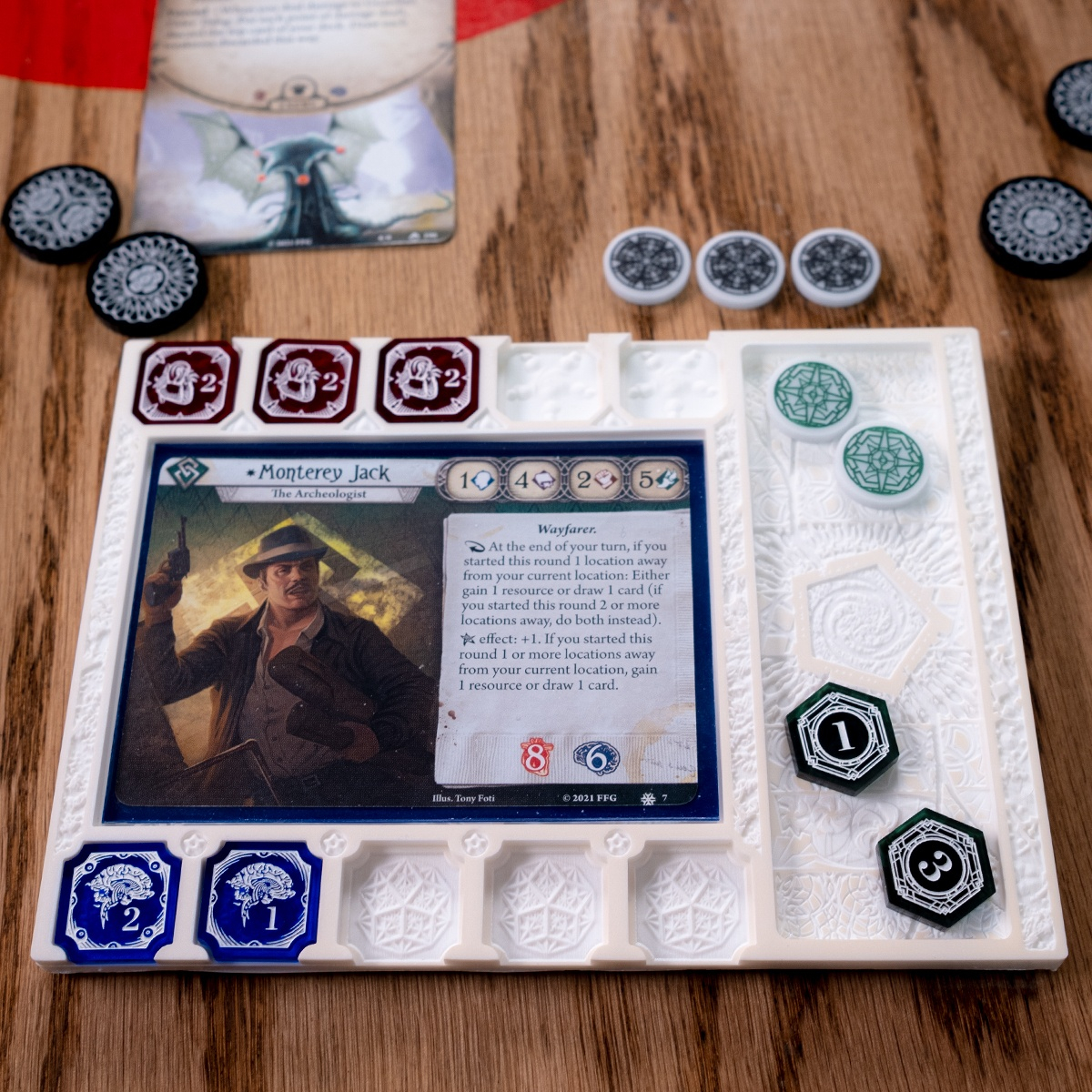 A straight on shot of the Edge of the Earth Mythos Board set up as it would be during a game of Arkham Horror LCG.  The investigator Monterey Jack is in the card holder spot with three two-value Damage Tokens above and two Sanity Tokens below, one two-value, and one one-value.  The board is surrounded by Arkham Horror LCG cards and Edge of the Earth Clue and Doom tokens