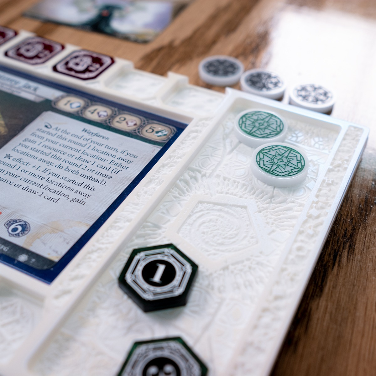 Close up of the Clue/Resource holder section of the Edge of Earth Mythos Board with minimal lighting, displaying the more neutral color of the white board, below two Resource Tokens and two Clue Tokens, designs inspired by Lovecraft’s The Mountains of Madness can be seen