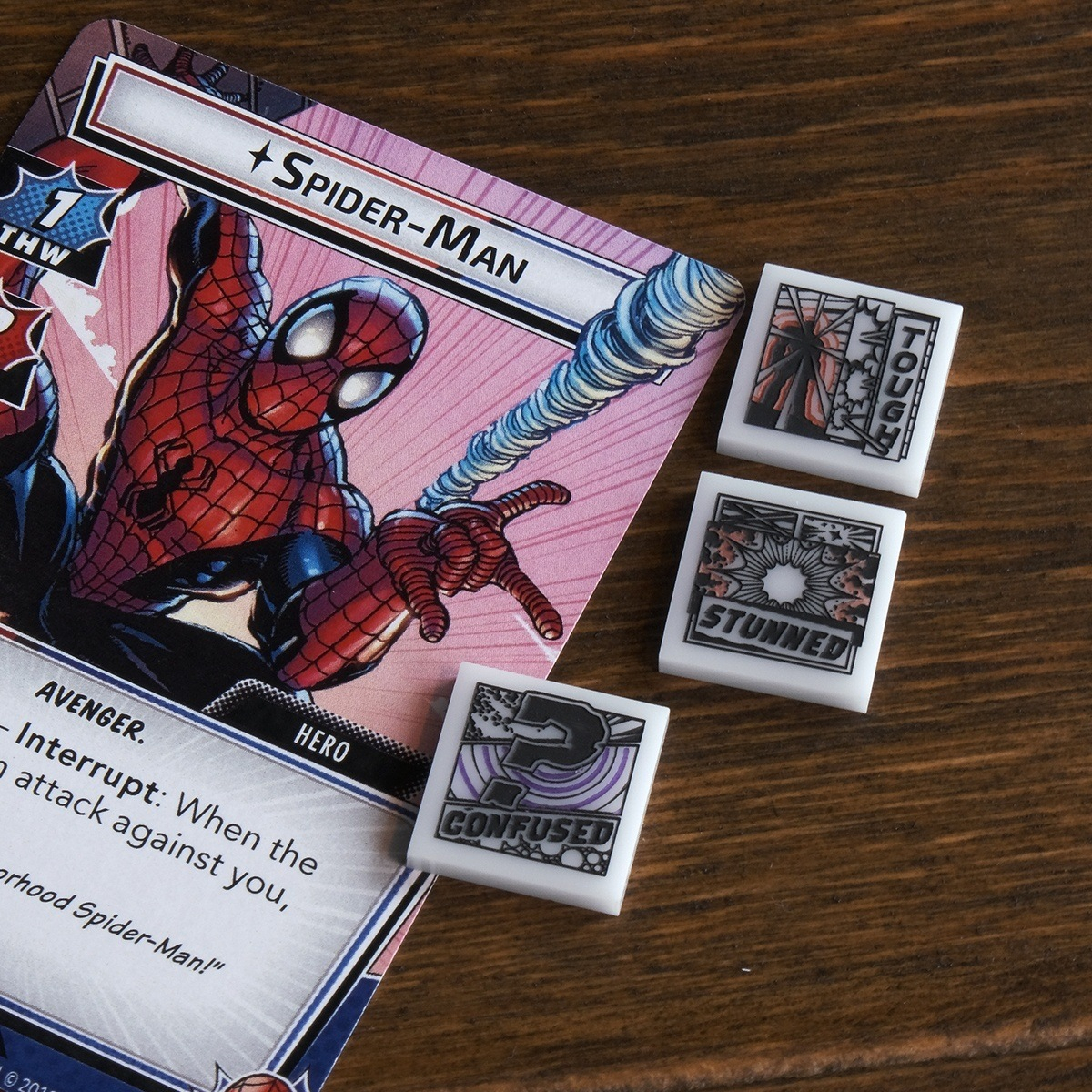 A Tough token, Stunned token, and Confused token are displayed on a wooden table just to the right of the Marvel Champions LCG card, Spider-Man
