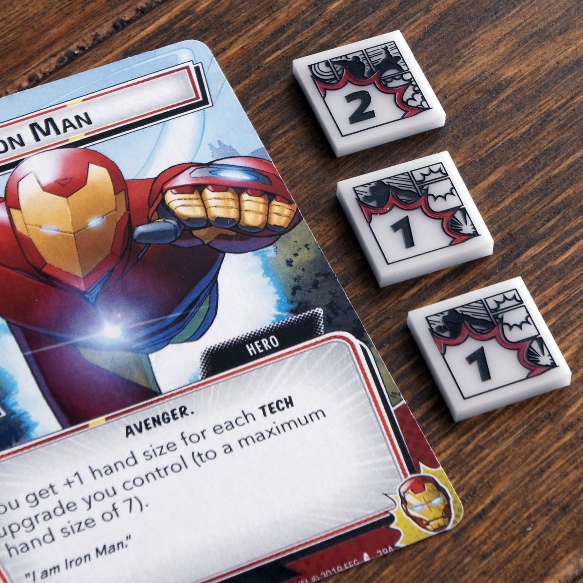 Three Damage tokens from the Hero Cosmic Tokens set are displayed on a wooden table just to the right of the Marvel Champions LCG card, Iron Man, one token is flipped to its two-damage side, the other two are on their one-damage side