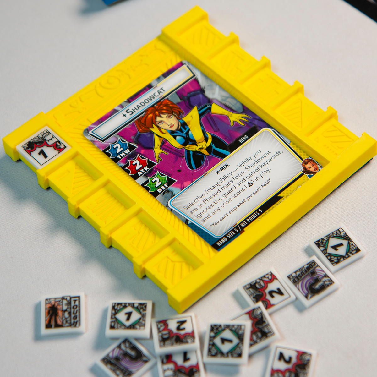 Yellow Beam Cosmic Board holding the Marvel LCG hero card, Shadowcat and a One Damage token.  Various tokens from the Hero Cosmic Token set surround the board