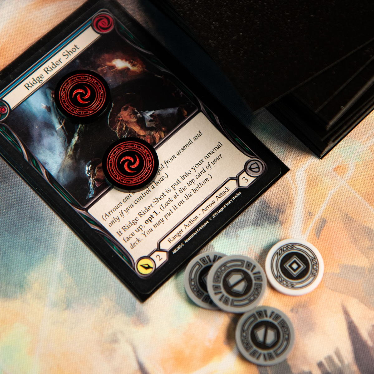 Two Resource Tokens on top of the Flesh and Blood TCG card, Ridge Rider Shot (Blue), three Armor Tokens and one Activation Token are below