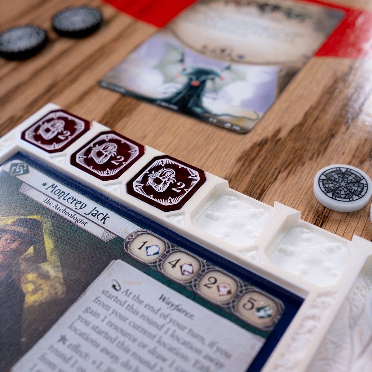 Close up of the Edge of Earth Mythos Board with three two-value Damage Tokens in the Damage slot, the empty Damage slots displaying designs of monstrous star-mounded graves and triangular striated markings.  Above the board is the Arkham Horror LCG Card, Guardian Elder Thing as well as some special edition Edge of the Earth Resource and Doom tokens
