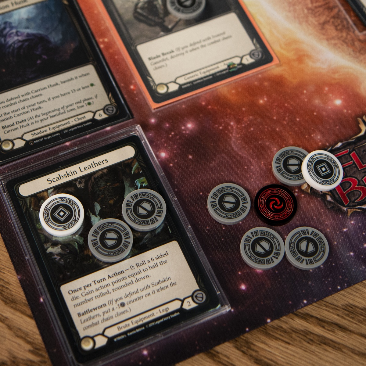 Flesh and Blood TCG equipment cards laid out for gameplay with one Activation Token and two Armor Tokens on the card Scabskin Leathers. An assortment of additional Majestic Tokens waits to be used nearby.