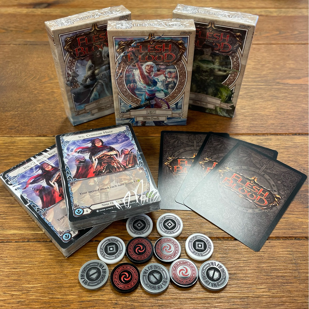 Oldhim, Lexi, and Briar Aria Blitz Decks, two Ira Decks, three Flesh and Blood TCG card backs denoting three random Flesh and Blood TCG promos, and a full set of Majestic tokens on a wooden table
