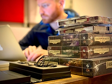 Unopened Arkham Horror products stacked on top of each other with Steven working on a laptop in the background.