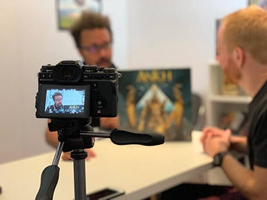 A camera's viewfinder in the foreground recording a conversation about the board game Ankh between Steven and Eric Lang in the background.