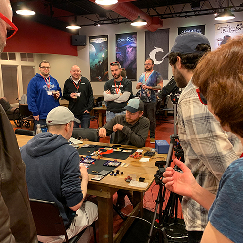Players standing around a table at Covenant watching Steven play against another player in Star Wars Destiny.